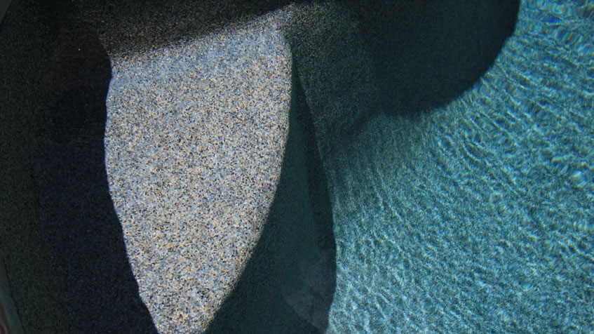 Pool step detail showing light-colored aggregate finish on the step tread with darker shadowed area and turquoise water visible to the right.