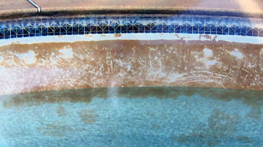 Pool wall showing significant plaster deterioration with large areas of brown discoloration and exposed substrate beneath, with blue decorative tile border at top and water visible at bottom.