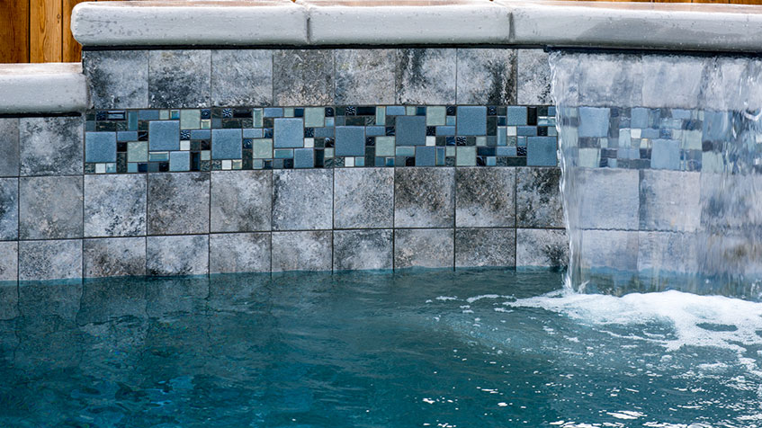 Pool spillway wall featuring decorative mosaic tile band in mixed shades of blue creating a pixelated pattern, with gray stone tiles above and below, and water flowing over the edge.