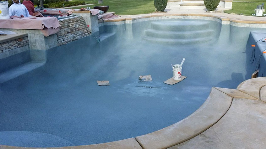 Pool under construction or resurfacing showing blue-coated interior surface with construction materials and tools scattered on the bottom, stone coping visible around edges.