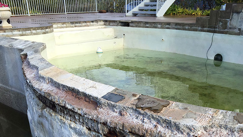 Deteriorating circular spa or pool showing green algae-filled water, damaged plaster and coping with exposed rebar, surrounded by concrete decking and metal fence.