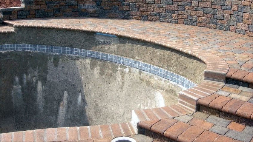 Empty circular pool showing weathered gray concrete interior with blue tile trim band, surrounded by decorative brick paver coping and patio with curved stone retaining wall in background.