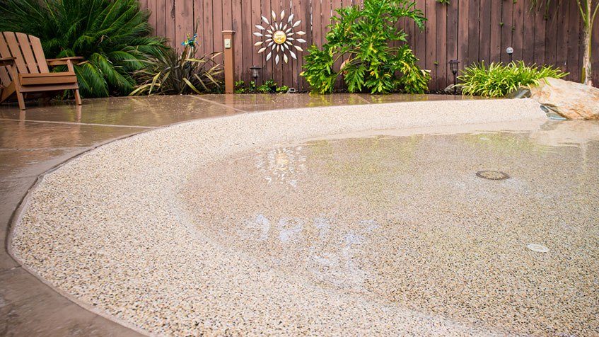 Wet pebble-finished pool deck or shallow water feature with cream-colored aggregate surface, tropical plants and wooden fence with decorative sun ornament visible in the background.