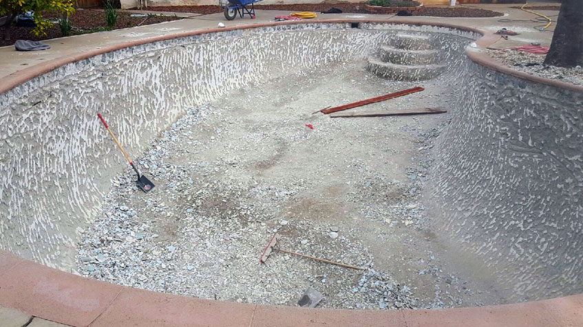 An empty kidney-shaped swimming pool under construction or renovation showing the gray plaster shell, with visible plumbing fixtures and built-in steps, surrounded by concrete decking.