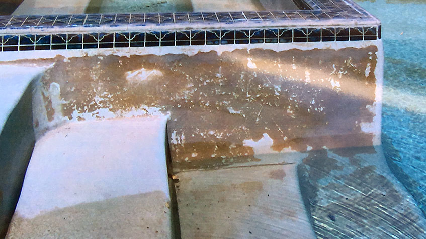 Deteriorating pool surface showing damaged plaster with extensive brown staining and exposed substrate, with blue and white decorative tile trim visible at the top edge.