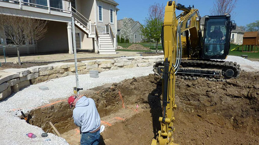 Backyard pool construction site with a yellow excavator digging near a house, showing excavated dirt area, gravel base, stone retaining wall, and a two-story house with deck in the background.