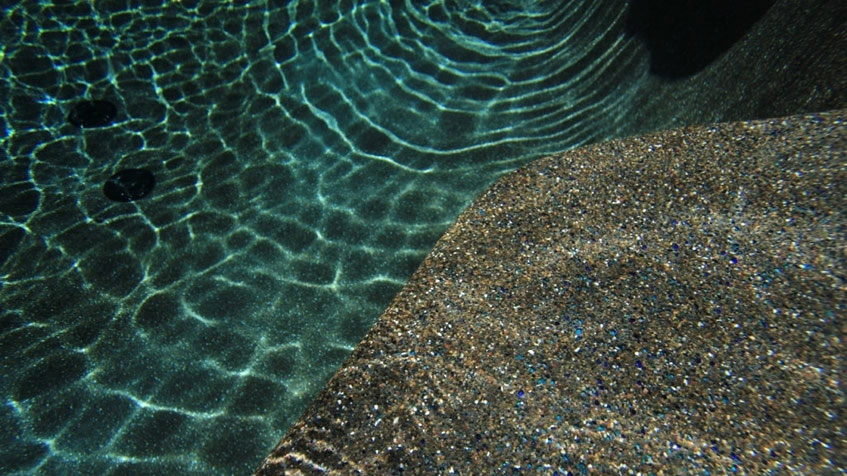 Underwater view of pool showing turquoise water with light reflections creating wavy patterns on the textured gray pebble-finished pool surface.