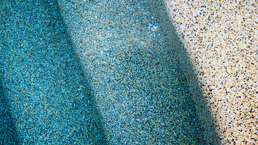 Close-up comparison of pool surface finishes showing blue pebble texture transitioning to beige/tan pebble texture, demonstrating different color options for pool plaster.