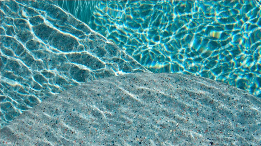 Underwater pool view showing sunlight creating rippling light patterns on gray pebble-textured pool surface through clear turquoise water.