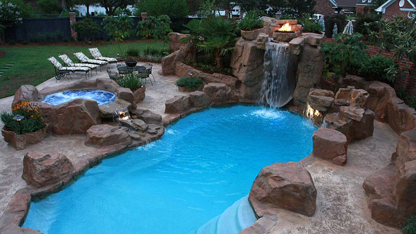 Resort-style backyard pool with bright blue water featuring large decorative boulders, a tall rock waterfall with fire feature on top, circular spa, stone patio with white lounge chairs, and residential houses visible in the background at dusk.