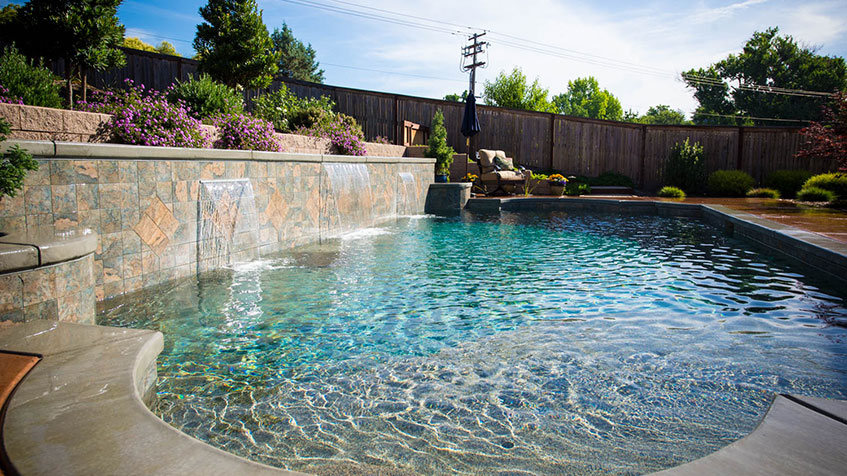 Swimming pool with stone waterfall wall featuring multiple water spouts, clear blue water, decorative plants on raised planter, wooden fence background, power lines visible above.