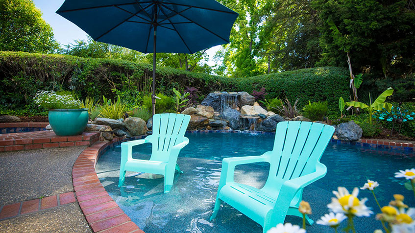 Small kidney-shaped pool with two turquoise Adirondack chairs placed in shallow water, blue patio umbrella, natural rock waterfall feature, lush tropical landscaping on terraced hillside, brick coping edge.