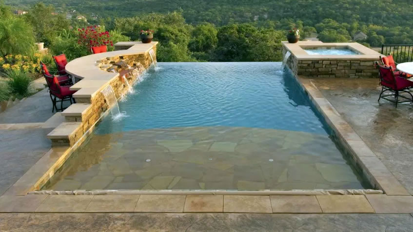 Infinity edge pool with stone wall and water spillways, overlooking forested valley view, stone patio with red lounge chairs, integrated spa area, natural stone coping and decking.