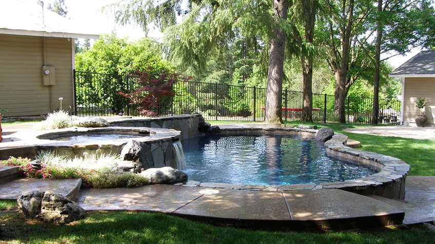 Raised perimeter overflow pool with dark interior, water flowing over all edges into catch basin, natural stone coping, metal fence, mature trees, residential setting with artificial turf and planted areas.