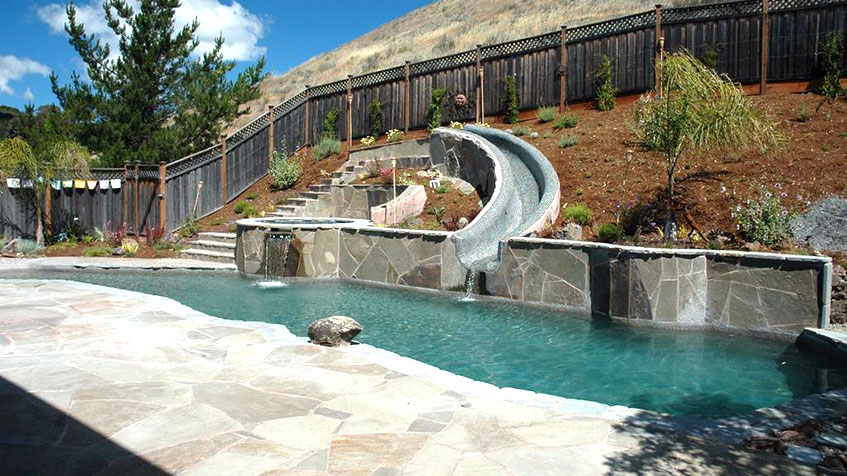 Resort-style backyard pool with bright blue water featuring large decorative boulders, a tall rock waterfall with fire feature on top, circular spa, stone patio with white lounge chairs, and residential houses visible in the background at dusk.