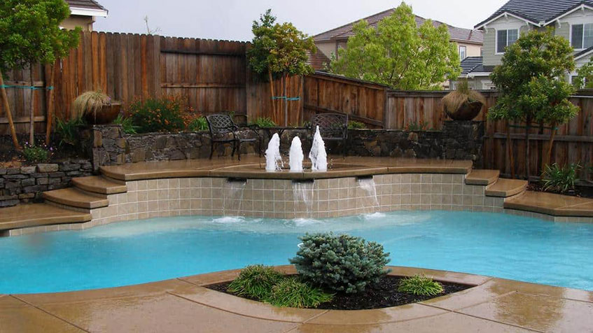 Backyard swimming pool with blue water featuring three decorative water fountains, raised spa area with stone wall, brown composite decking steps, wooden privacy fence, circular planted island in foreground, and neighboring houses visible.
