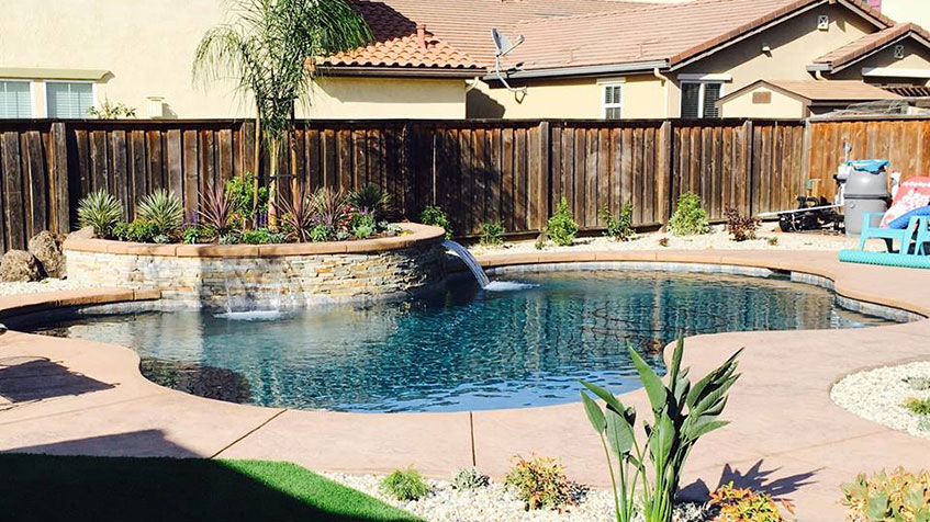 Freeform swimming pool with blue water, raised curved planter with stone waterfall feature, tan concrete decking, wooden fence, residential house with tile roof in background, and colorful pool toys visible on deck.