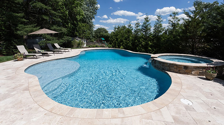 Large freeform pool with clear blue water, circular shallow sun shelf area, elevated round spa with stone sides, white stone patio decking, lounge chairs with umbrella, surrounded by mature trees.
