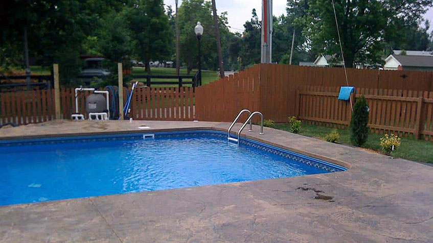 Rectangular swimming pool with blue water, metal ladder, brown stamped concrete decking, wooden privacy fence, residential neighborhood setting with trees and utility poles visible.
