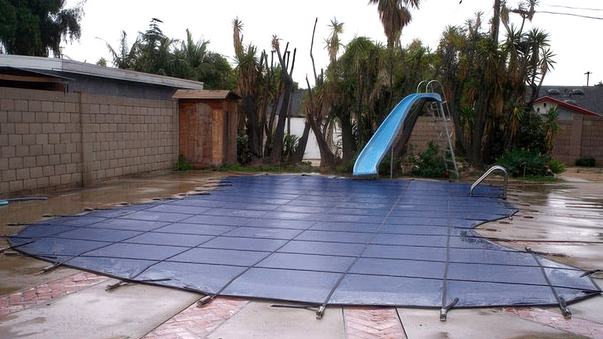 Covered swimming pool with gray safety cover secured by straps, blue water slide in background, wet concrete deck, concrete block wall, palm trees and residential buildings visible.